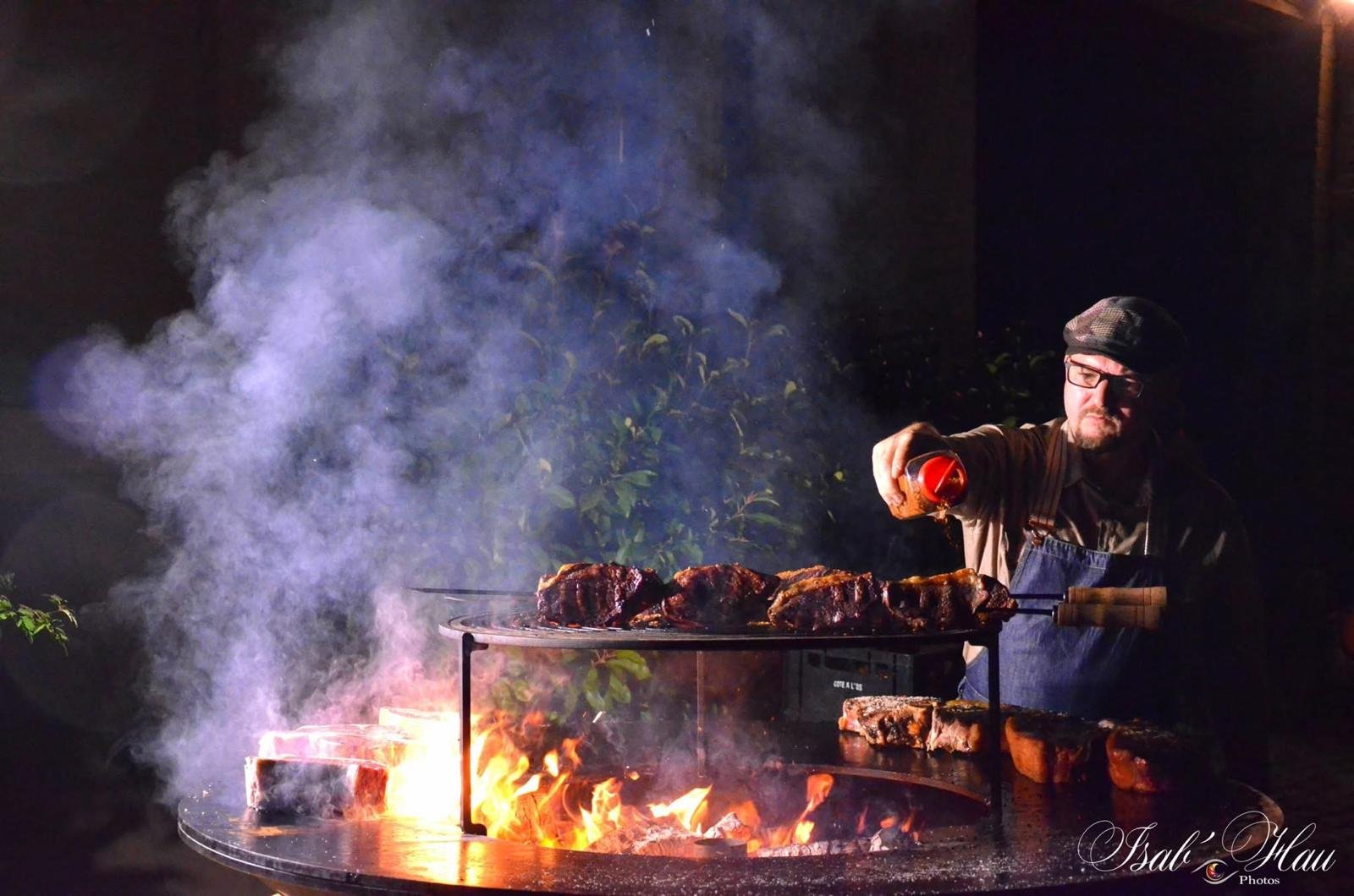 Un homme portant des lunettes et une casquette fait griller de gros morceaux de viande sur une flamme nue, la nuit, en versant un liquide rouge sur les aliments. La fumée s'élève autour du gril, créant une atmosphère dramatique et enfumée.